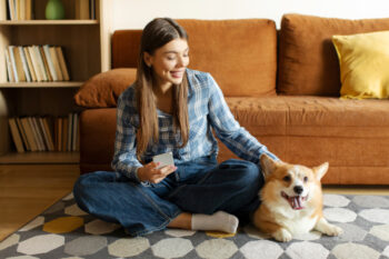 Best friend concept. Happy woman holding cellphone and petting her Welsh Corgi dog, sitting on floor carpet in living room